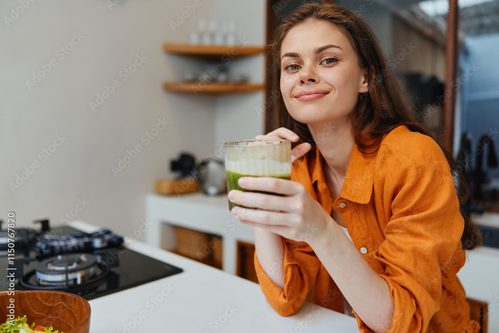 Young woman with long brown hair smiles while holding a green smoothie in a modern kitchen, wearing an orange shirt Healthy lifestyle and nutrition concept Young woman with long brown hair smiles while holding a green smoothie in a modern kitchen, wearing an orange shirt Healthy lifestyle and nutrition concept