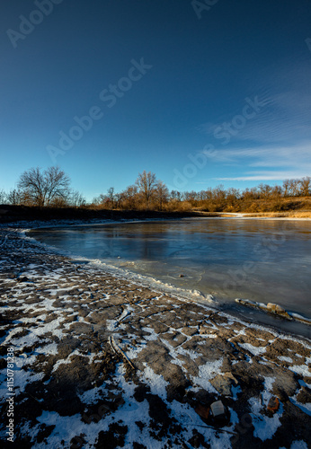 Feozen pond at the winter day,lake with ice,dry ground near the lake.Sun in the sky, sunlights and sky with blue clouds.Blue winter colors , wild nature with a pond.Frozen beach near the lake,snowy  photo