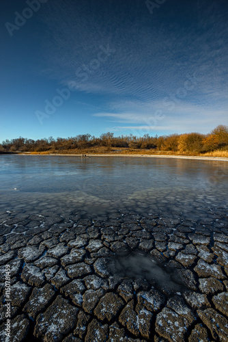 Feozen pond at the winter day,lake with ice,dry ground near the lake.Sun in the sky, sunlights and sky with blue clouds.Blue winter colors , wild nature with a pond.Frozen beach near the lake,snowy  photo