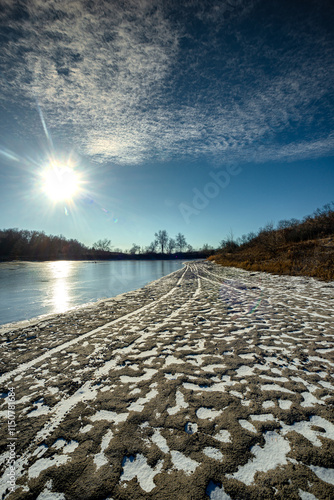 Feozen pond at the winter day,lake with ice,dry ground near the lake.Sun in the sky, sunlights and sky with blue clouds.Blue winter colors , wild nature with a pond.Frozen beach near the lake,snowy  photo