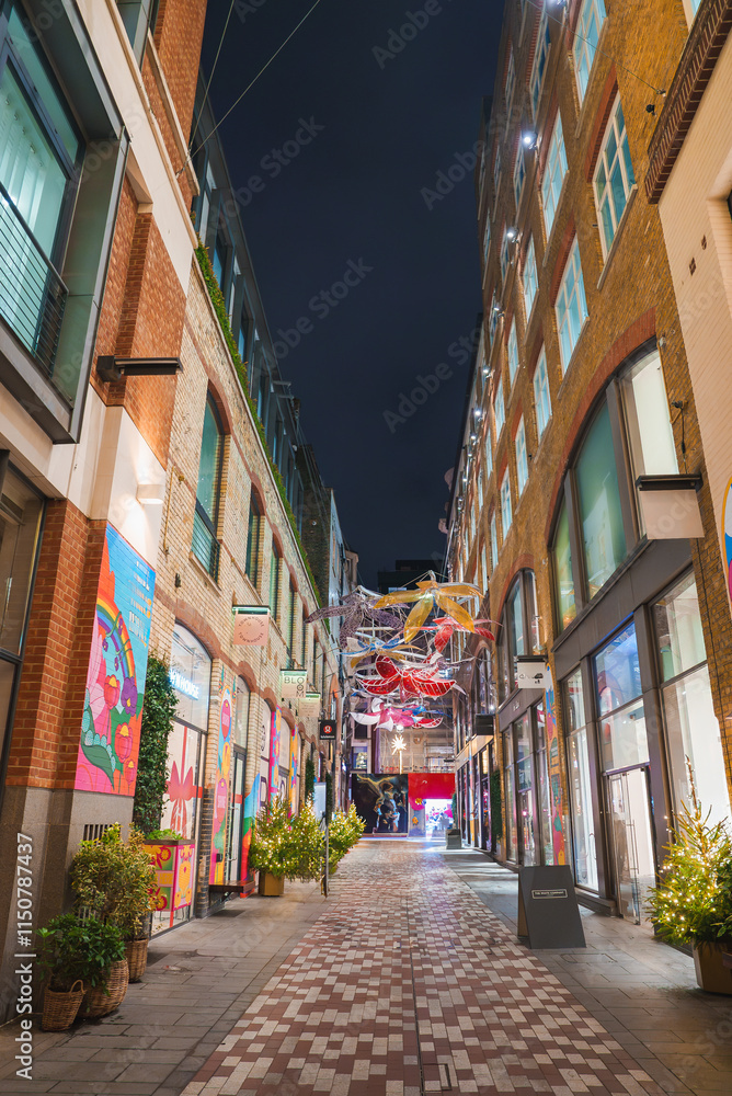 Fototapeta premium A vibrant London street features festive Christmas lights, tall brick buildings, colorful murals, and potted plants, creating a lively holiday atmosphere.