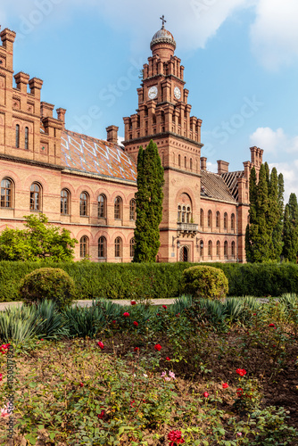 Canvas Print A large brick building with a clock tower and a courtyard with flowers