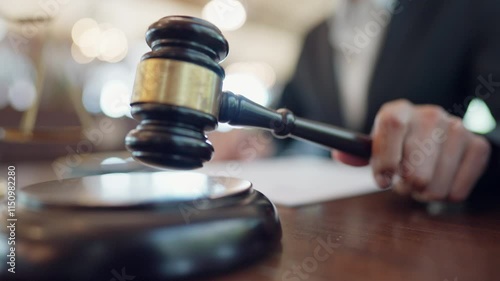 A man is holding a gavel and writing on a piece of paper. The gavel is on a wooden surface and the man is wearing a suit. Scene is serious and formal
