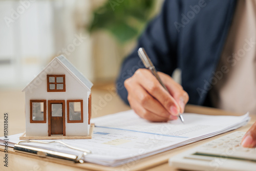 A man writes a signature on house insurance documents, using a calculator for tax and property details. Counting payments and rent by model house, he finalizes the price photo