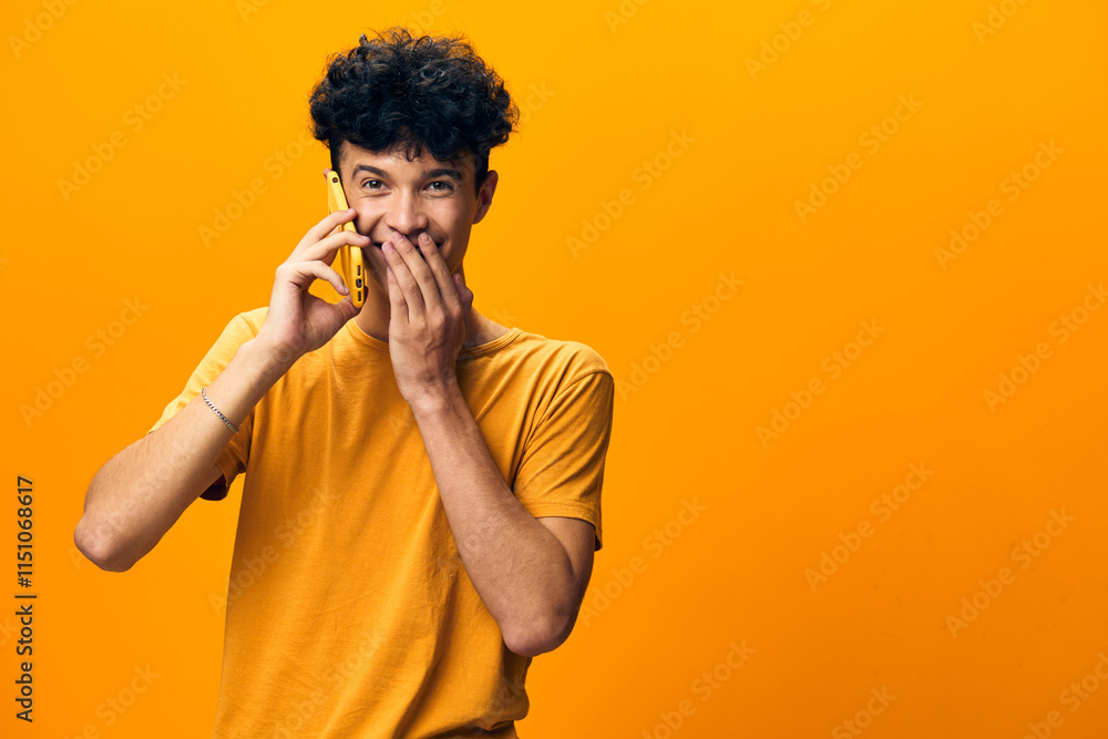 Young man smiling while talking on a yellow phone against a vibrant orange background, expressing joy and excitement Young man smiling while talking on a yellow phone against a vibrant orange background, expressing joy and excitement