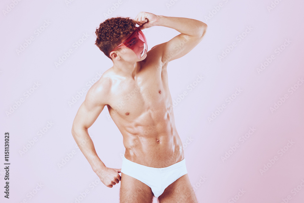Obraz premium Confident male model posing in white swimwear, wearing stylish pink sunglasses, showcasing fitness and fashion. Trendy summer look with a playful vibe.