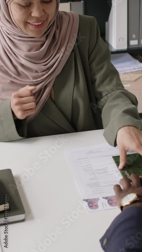 Vertical tilt shot of happy Muslim woman in hijab getting her visa approved, Black male visa officer sealing her application during interview in consulate