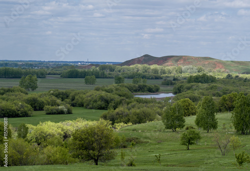 A river valley covered with trees and shrubs against a backdrop of low mountains. The sun breaks through the clouds.