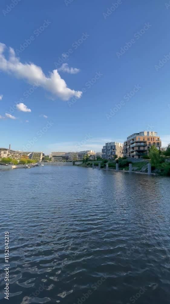 A scenic riverside view featuring modern architecture, lush greenery, and a tranquil blue sky dotted with clouds. The rippling water adds to the peaceful charm of the urban setting.