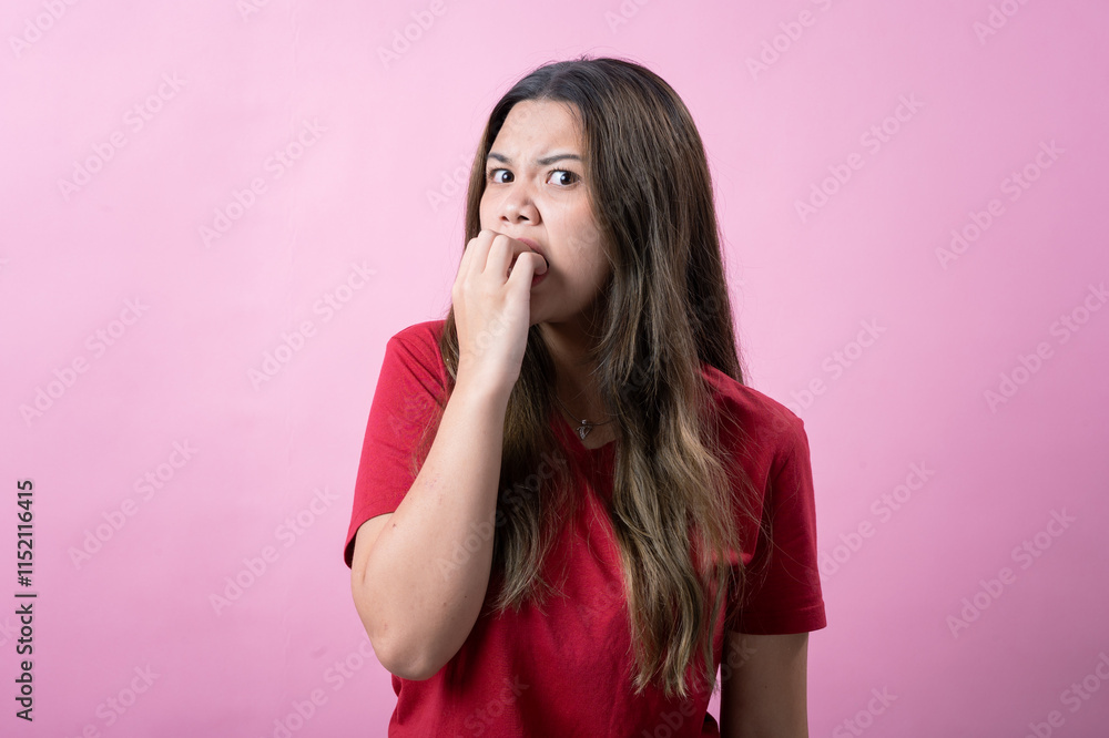 Asian woman in a red t-shirt standing against a pink background, showing a worried or nervous expression while biting her nails and frowning, depicting stress, anxiety Asian woman in a red t-shirt standing against a pink background, showing a worried or nervous expression while biting her nails and frowning, depicting stress, anxiety