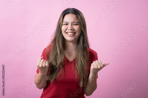 A happy young woman with long brown hair and light skin, wearing a red T-shirt, smiles brightly and clenches her fists in a celebratory and enthusiastic gesture against a pink background. photo