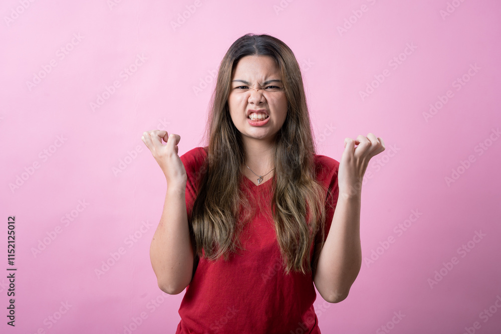Frustrated young woman in a red shirt makes a confused gesture with both hands, showing irritation against a pink background. The playful and exaggerated facial expression adds humor and personality.