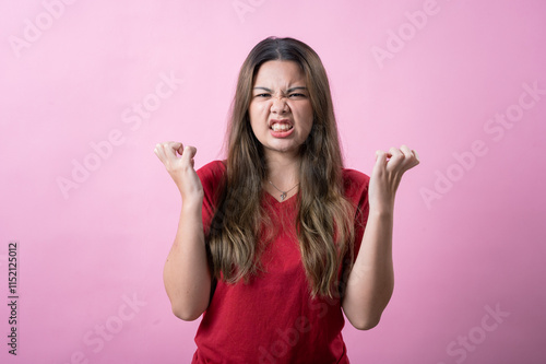 Frustrated young woman in a red shirt makes a confused gesture with both hands, showing irritation against a pink background. The playful and exaggerated facial expression adds humor and personality.