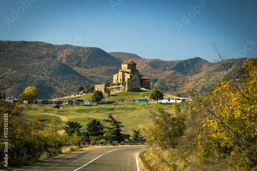 Jvari Monastery perched on a hill, surrounded by autumn-colored mountains under a clear blue sky. A stunning blend of historic architecture and vibrant seasonal landscapes photo