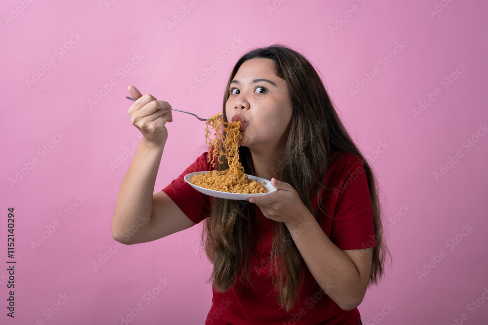 Asian woman in a red shirt enjoys eating noodles from a white plate using a fork, smiling playfully and looking at the camera against a solid pink background. Asian woman in a red shirt enjoys eating noodles from a white plate using a fork, smiling playfully and looking at the camera against a solid pink background.