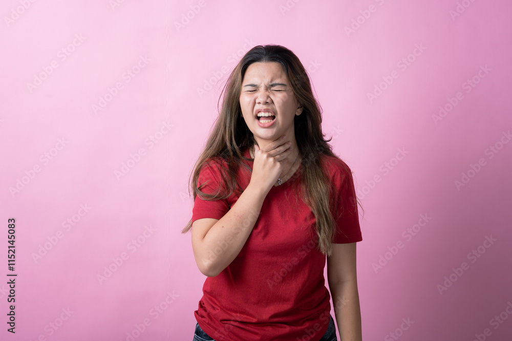 Asian woman in a red shirt with long brown hair shows discomfort, grimacing and clutching her throat with one hand against a pink background. Asian woman in a red shirt with long brown hair shows discomfort, grimacing and clutching her throat with one hand against a pink background.