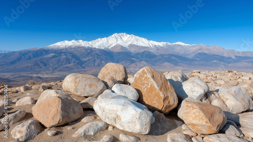 A rugged desert landscape with large boulders in the foreground, framed by majestic snow-capped mountains under a vivid blue sky. photo