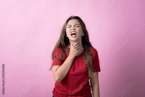 Fotografi Asian woman in a red shirt with long brown hair shows discomfort, grimacing and clutching her throat with one hand against a pink background