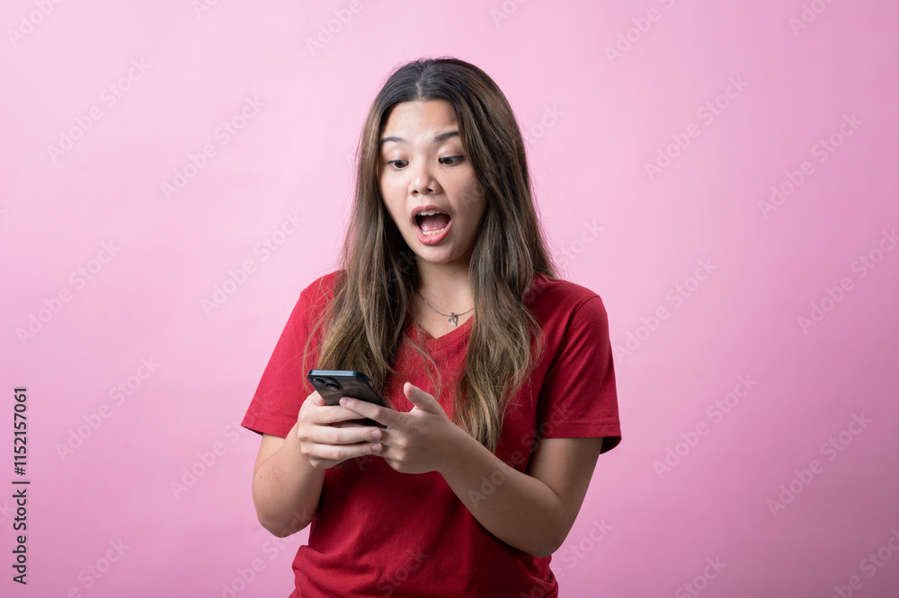 Surprised young woman with long brown hair wearing a red t-shirt looks at her smartphone against a pink background. Her shocked expression conveys excitement, disbelief, or unexpected news. Surprised young woman with long brown hair wearing a red t-shirt looks at her smartphone against a pink background. Her shocked expression conveys excitement, disbelief, or unexpected news.