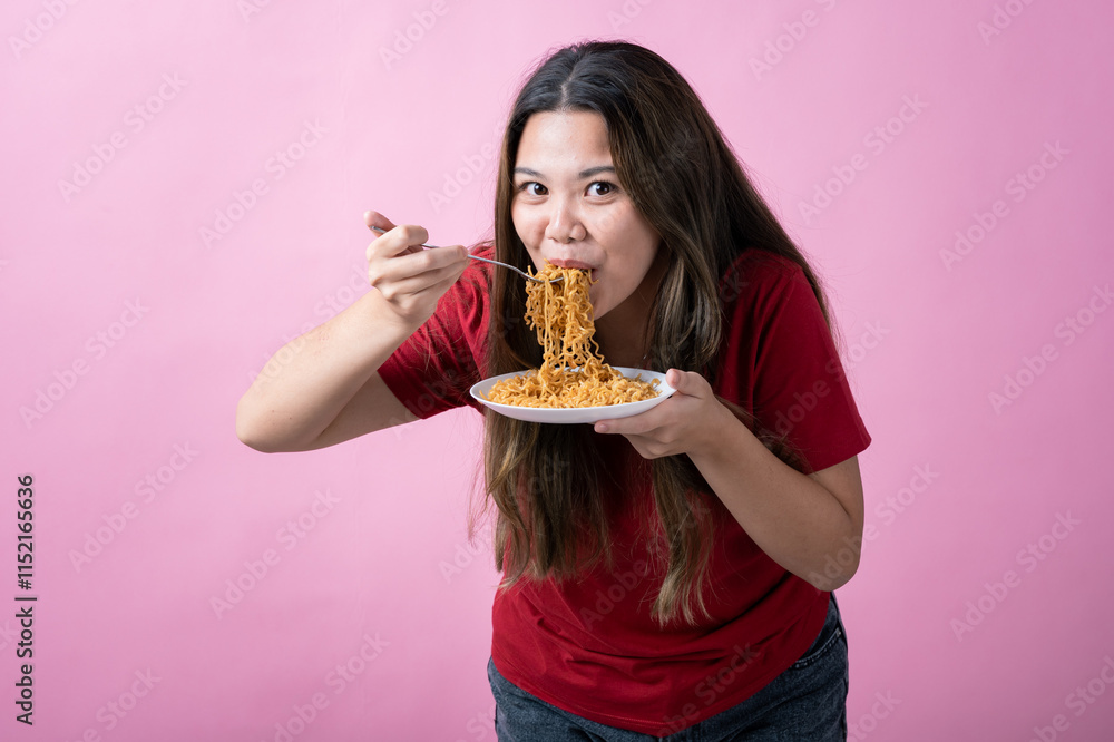 Asian woman in a red shirt with long brown hair eagerly eating noodles from a white plate with a fork, against a solid pink background. Asian woman in a red shirt with long brown hair eagerly eating noodles from a white plate with a fork, against a solid pink background.