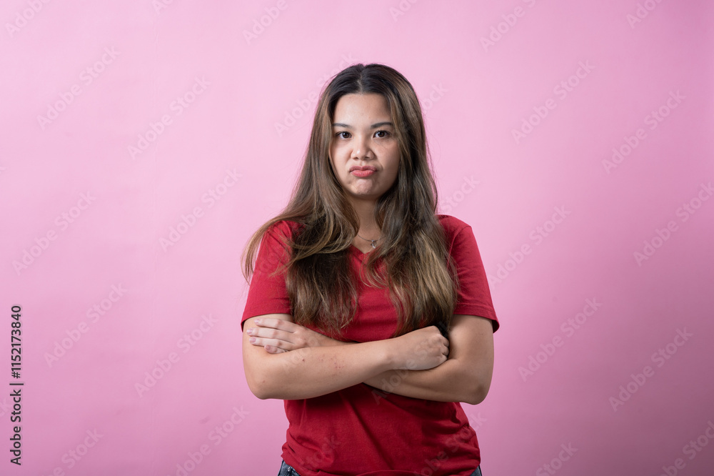 Asian woman in a red shirt with long brown hair crosses her arms and makes a pouty, dissatisfied expression against a vibrant pink background. Asian woman in a red shirt with long brown hair crosses her arms and makes a pouty, dissatisfied expression against a vibrant pink background.