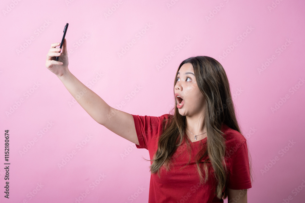 Young woman with long brown hair in a red t-shirt poses for a selfie, puckering lips for a playful expression against a pink background. She holds a smartphone, capturing a fun and casual moment. Young woman with long brown hair in a red t-shirt poses for a selfie, puckering lips for a playful expression against a pink background. She holds a smartphone, capturing a fun and casual moment.