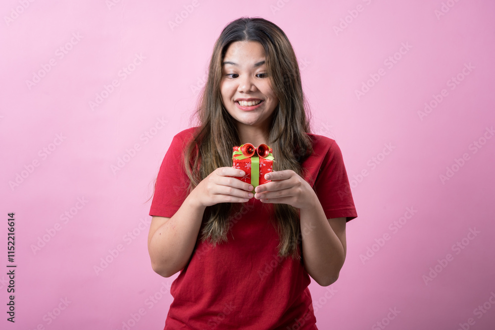 A cheerful young woman with long brown hair and light skin, wearing a red T-shirt, holds a small red gift box with a green ribbon and a decorative bow, smiling brightly against a pink background. A cheerful young woman with long brown hair and light skin, wearing a red T-shirt, holds a small red gift box with a green ribbon and a decorative bow, smiling brightly against a pink background.