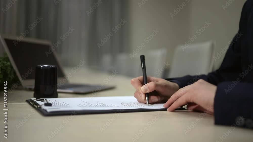 Anonymous person sitting at sleek desk in contemporary office while signing document with pen. Modern laptop and small plant are visible in the background. Anonymous person sitting at sleek desk in contemporary office while signing document with pen. Modern laptop and small plant are visible in the background.