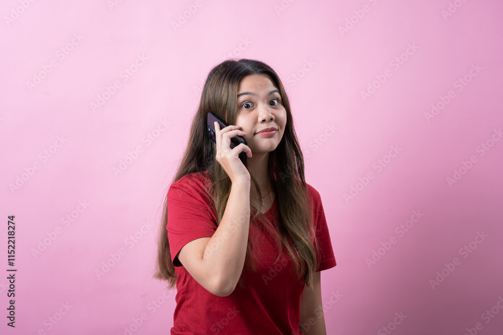 Surprised young woman with long brown hair wearing a red t-shirt talks on a smartphone against a pink background, reacting with shock or excitement during a phone call, expressing strong emotions. Surprised young woman with long brown hair wearing a red t-shirt talks on a smartphone against a pink background, reacting with shock or excitement during a phone call, expressing strong emotions.