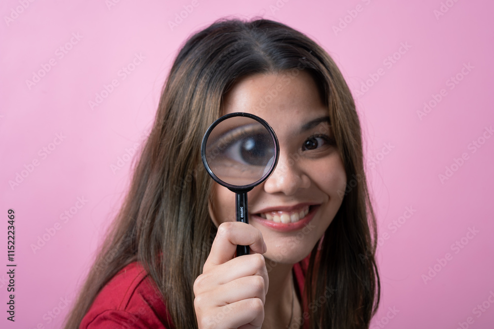 A young woman with long brown hair , wearing a red T-shirt, smiles playfully while holding a magnifying glass up to her eye, enlarging it for a fun and curious effect, against a pink background. A young woman with long brown hair , wearing a red T-shirt, smiles playfully while holding a magnifying glass up to her eye, enlarging it for a fun and curious effect, against a pink background.