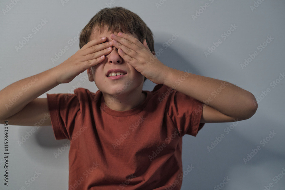 A boy covering his eyes with both hands, expressing a playful or thoughtful moment. The plain background emphasizes his pose and gesture. A boy covering his eyes with both hands, expressing a playful or thoughtful moment. The plain background emphasizes his pose and gesture.