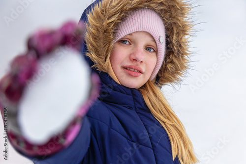 Girl in winter coat holding snowball forward. A young girl dressed in a navy blue winter coat with a fur-trimmed hood and a pink knit hat holds a snowball toward the camera. photo