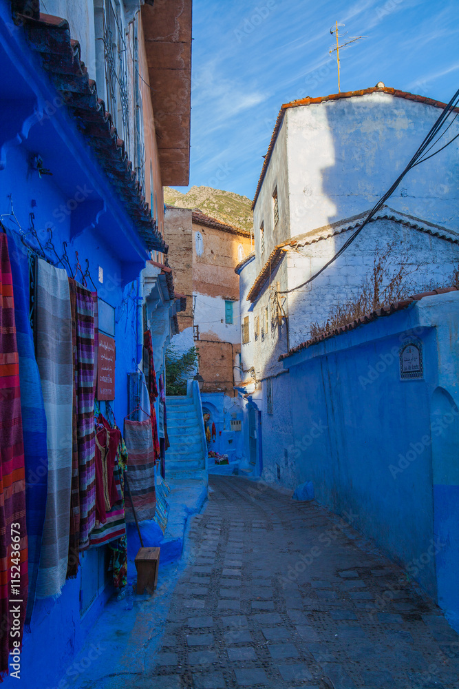 narrow, winding street in a blue-washed town, with colorful textiles displayed outside a building and a view of distant hills narrow, winding street in a blue-washed town, with colorful textiles displayed outside a building and a view of distant hills