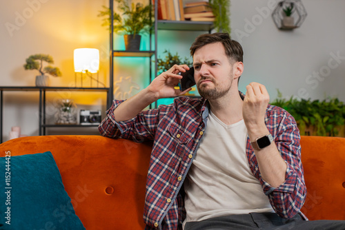 Angry young man arguing on smartphone while sitting on couch at home. Quarrel. Caucasian mad guy talking loudly and shouting into cellphone, maliciously conveying and proving point through gestures. photo