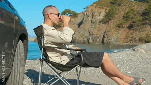 Man relaxing on beach drinking from metal cup. Tourist enjoys a hot drink while relaxing on a shingle beach beside a car. Admiring the ocean view and cliffs creates a peaceful and content atmosphere