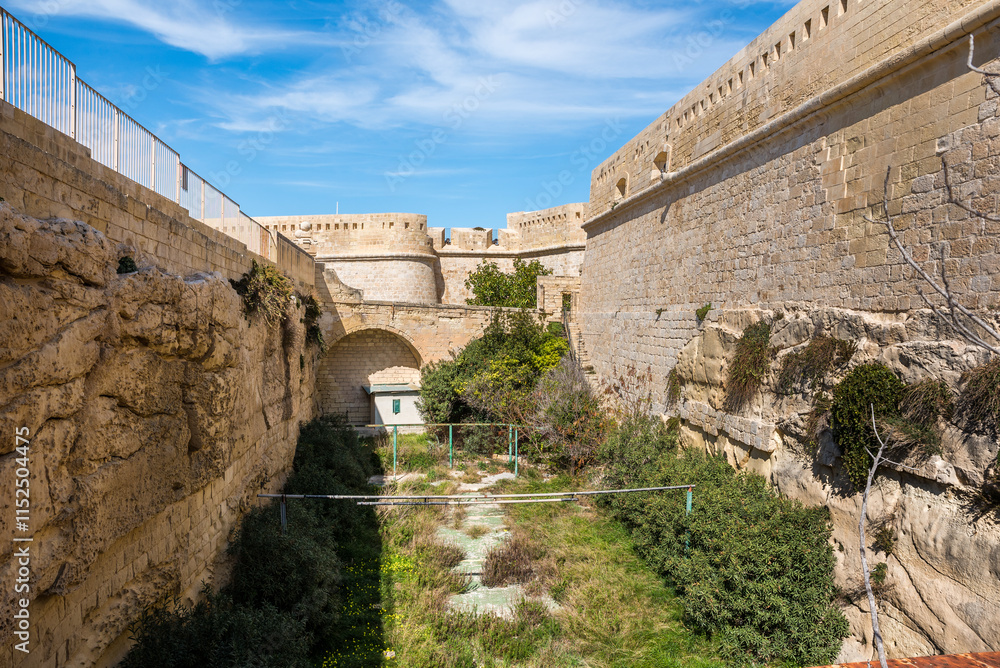 Fototapeta premium Historic city walls of Valletta, Malta, showcasing ancient fortifications, a landscaped walkway, and bright Mediterranean architecture.