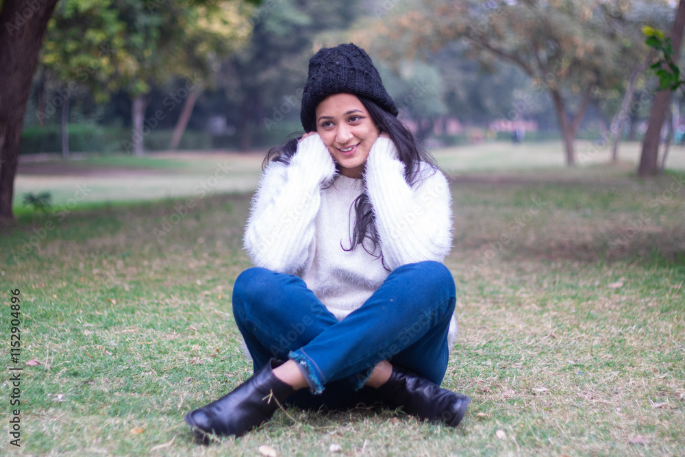 Young happy indian woman wearing warm white sweater and black woolen hat sitting outdoor at park having fun in winter season. Young happy indian woman wearing warm white sweater and black woolen hat sitting outdoor at park having fun in winter season.