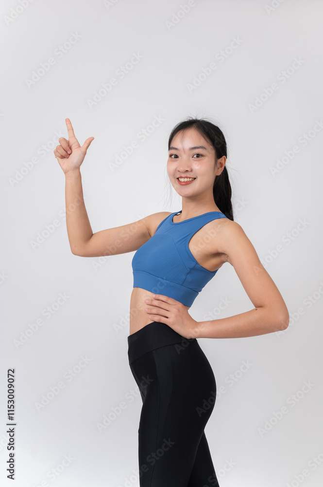 A confident, slim woman in sportswear pointing upward and smiling at the camera, studio background. A confident, slim woman in sportswear pointing upward and smiling at the camera, studio background.