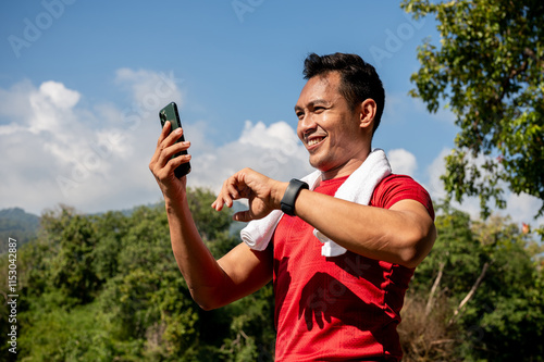 A happy, sporty Asian man in sportswear uses his smartphone while jogging in a park on a sunny day.
