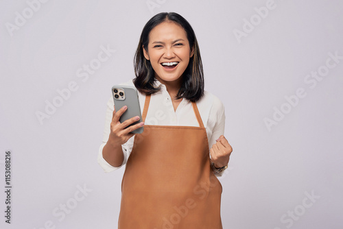 Smiling young Asian woman barista barman employee wearing a brown apron working in coffee shop holding mobile phone and celebrating success isolated on white background. Small business startup concept