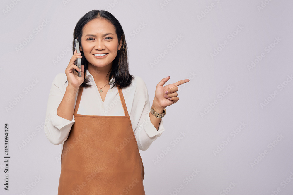 Smiling young Asian woman barista barman employee wearing a brown apron working in coffee shop talking on mobile phone and pointing finger at copy space isolated on white background Smiling young Asian woman barista barman employee wearing a brown apron working in coffee shop talking on mobile phone and pointing finger at copy space isolated on white background