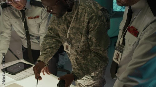 Medium tilt-up shot of African American male lieutenant in camouflage uniform briefing Caucasian male and female officers on enemy positions in command center, pointing to warfare maps on light table photo