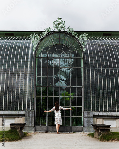 a woman stands with her back to the camera in front of a large glass door that is part of a greenhouse or greenhouse. The door has an arched shape and is decorated with decorative elements on top. photo