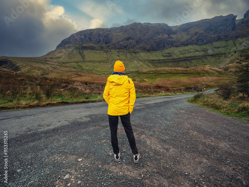 Travel and tourism in Ireland. Teenager girl standing on a side of a road and looking at majestic nature scenery of Gleniff Horseshoe drive with tall mountains. Model wearing yellow jacket and hat. photo