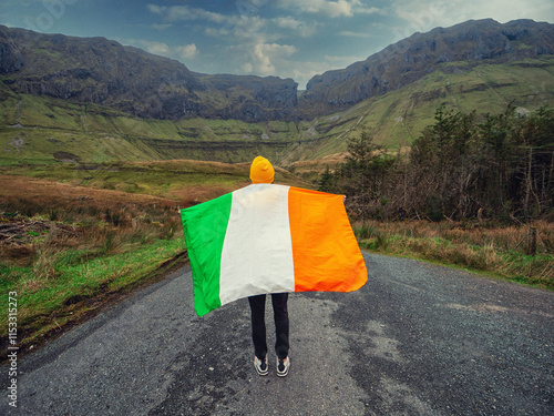 Travel and tourism in Ireland. Teenager girl with flag of Ireland on a road and looking at majestic nature scenery of Gleniff Horseshoe drive with tall mountains. Model wearing yellow jacket and hat. photo