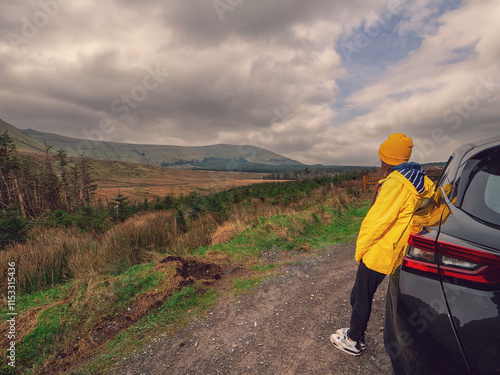 Travel and tourism in Ireland. Young woman standing by black car and looking at looking at majestic nature scenery of Gleniff Horseshoe drive with tall mountains. Model wearing yellow jacket and hat. photo