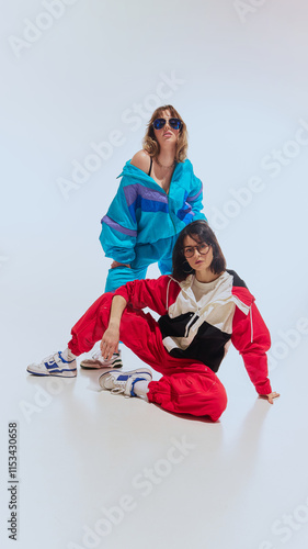 Obraz na płótnie Two women in bright, retro windbreakers, glasses and flashy tracksuits from the 90s, exuding confidence, posing against white studio background