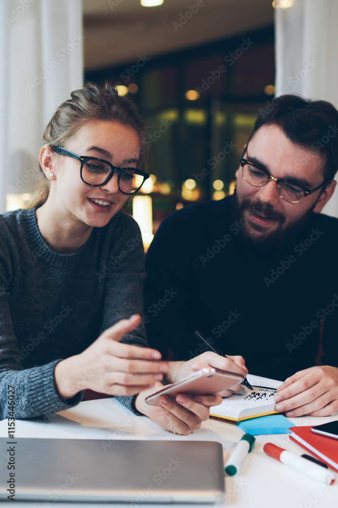 Fototapeta premium Smiling hipster girl holding mobile phone in hand feeling good from received funny message, woman using smartphone connected to 4g internet at university cafeteria spending time with boyfriend