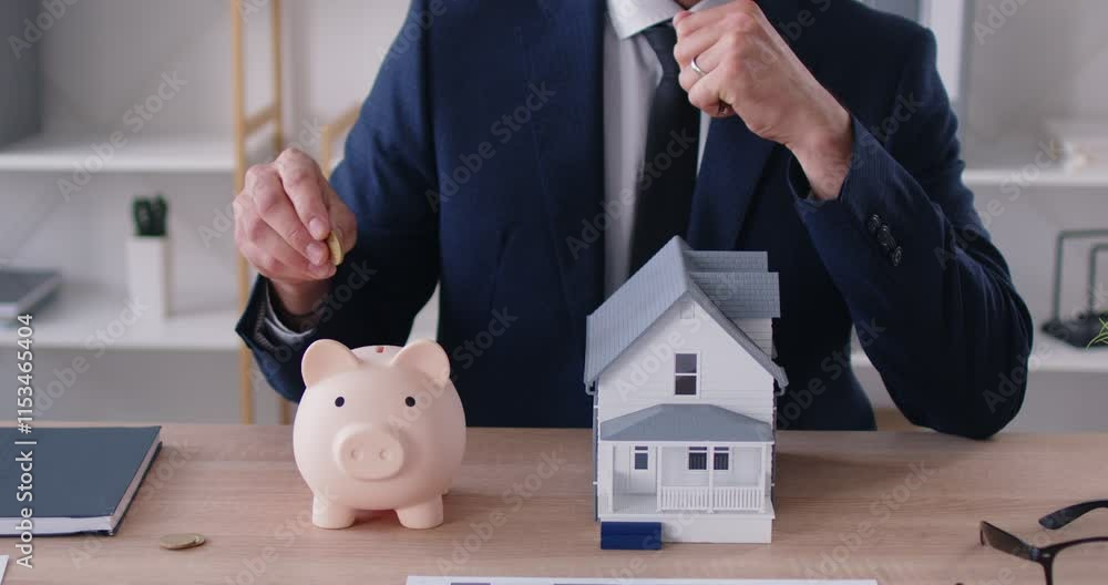 Man realtor in suit sitting at the desk with wooden toy house, money coins and piggy bank at table giving house keys to camera. Saving money for real estate. 4k video. slow motion video.
