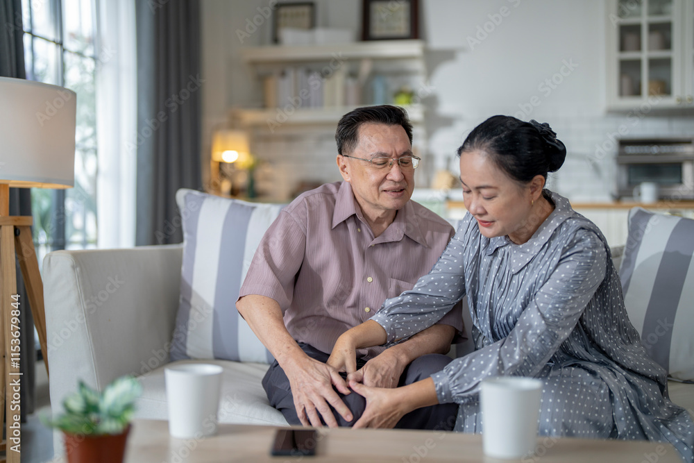 An elderly couple sits in their living room. The man holds his knee, appearing to be in discomfort, while his partner offers support. The image conveys care, concern, and the bond between the couple. An elderly couple sits in their living room. The man holds his knee, appearing to be in discomfort, while his partner offers support. The image conveys care, concern, and the bond between the couple.